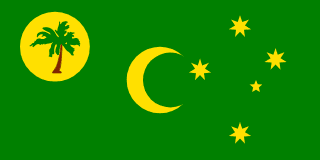 The flag of the Cocos (Keeling) Islands consists of a green field with a palm tree on a gold disc in the canton, a gold crescent moon in the center of the flag, and a gold southern cross in the fly side.