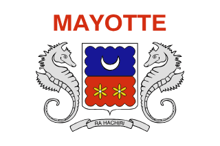 The flag of Mayotte bears a white field with the national coat of arms in the center and the country's name in red above it. The coat of arms consists of a shield supported by two seahorses and the national motto in a banner below it. The shield has a white fringe and bears a blue field with an upwards-facing white crescent on the top half and a red field with two yellow ylang-ylang flowers on the bottom half.